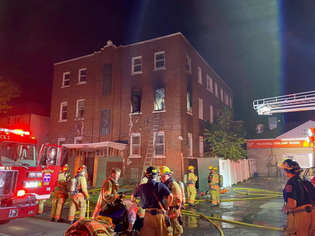 a view of the northeast corner of the building. A second-floor apartment, damaged by fire, can be seen. Fire crews can be seen in the foreground, regrouping after bringing the blaze under control.